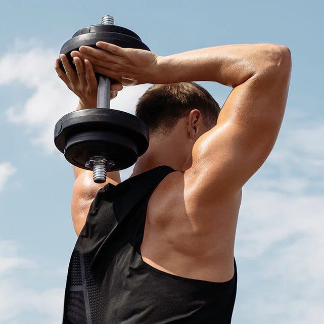 Athletic man performing an overhead dumbbell triceps extension outdoors, showcasing toned back, shoulder, and arm muscles against a clear blue sky background.