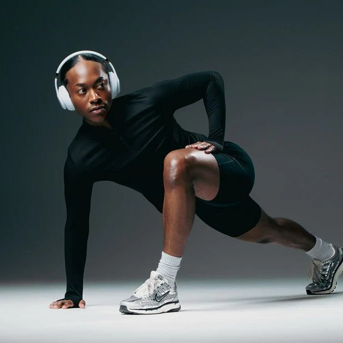 Athlete performing a deep side lunge stretch while wearing wireless headphones, black activewear, and running shoes in a modern fitness studio with a dark background.
